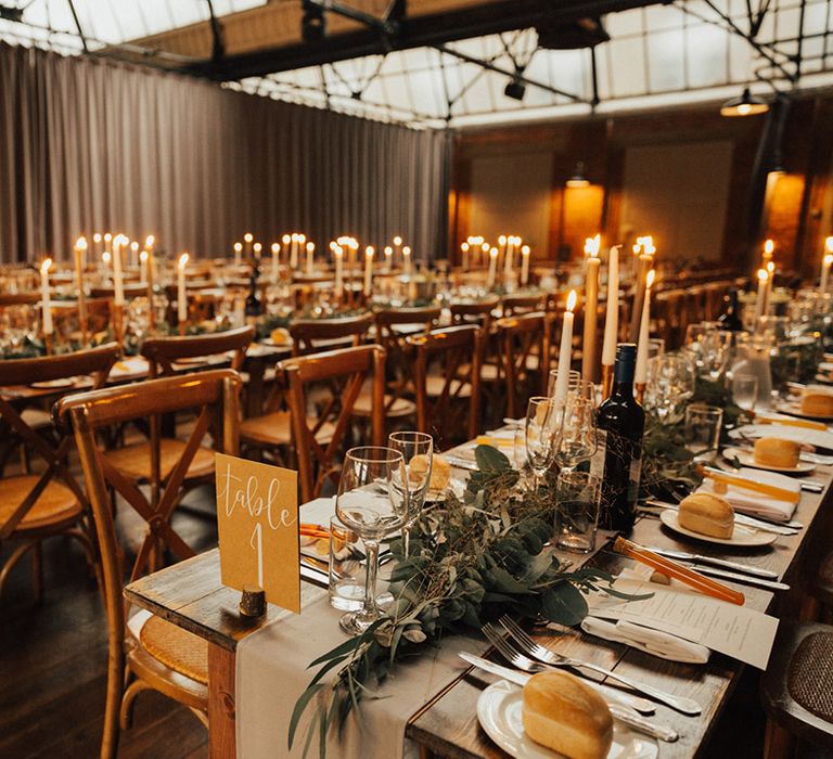 Wooden banquet tables with neutral fabric table runners and green foliage complete with candlesticks 