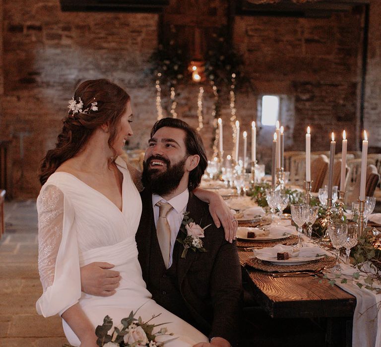 Groom in black tie seated at the table for the reception with the bride on his lap in a long sleeve wedding dress holding a pink rose bouquet 