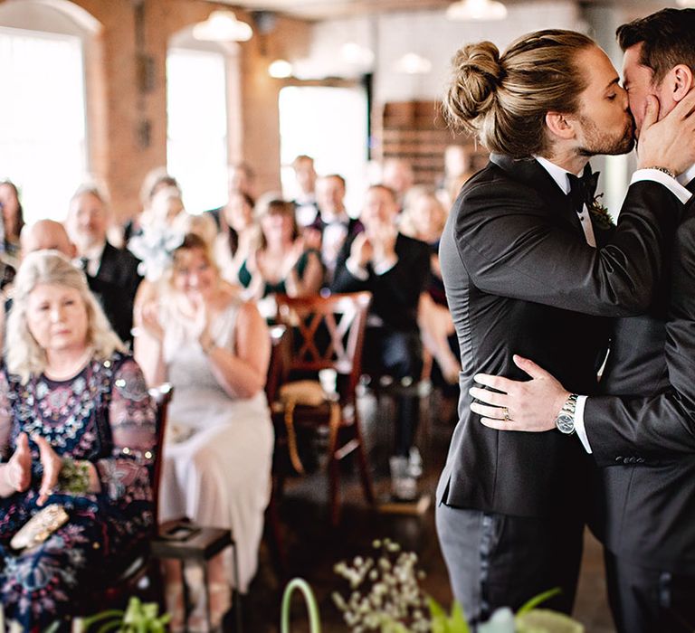 Grooms in black-tie kiss during botanical wedding ceremony with industrial vibe 