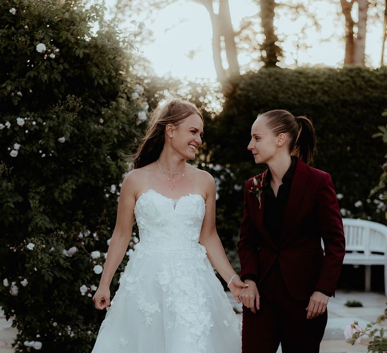 Bride in a strapless flower wedding dress walking with the bride in a burgundy suit and black shirt for their rustic wedding