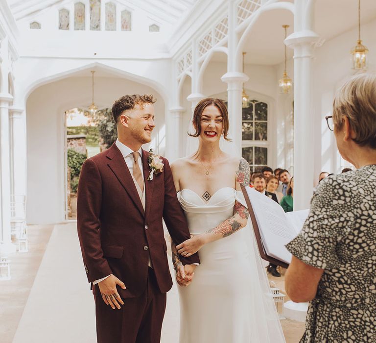 Bride in Vera Wang wedding dress stands with her groom in maroon suit on during wedding ceremony