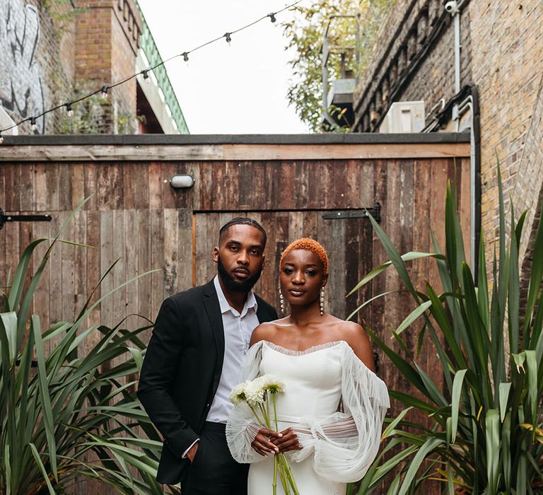 Bride in off shoulder wedding dress with long lace puff sleeves holding white dahlias standing with groom in skinny black tux at 100 Barrington wedding venue