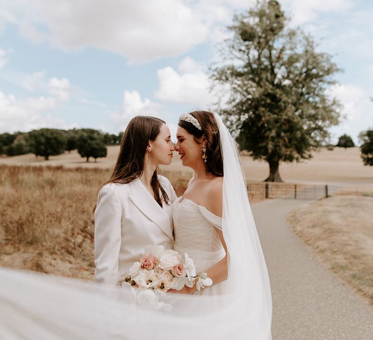 Bride in white suit smiling at the bride in an off the shoulder wedding dress holding a pale pink and white rose and orchid bouquet 