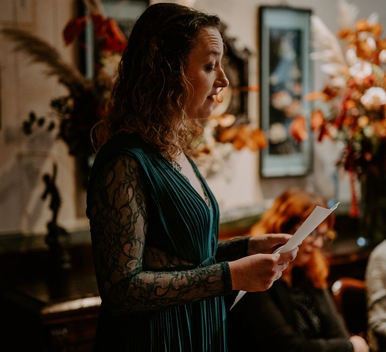 Bridesmaid in bottle green bridesmaid dress with long lace sleeves doing reading at autumnal wedding