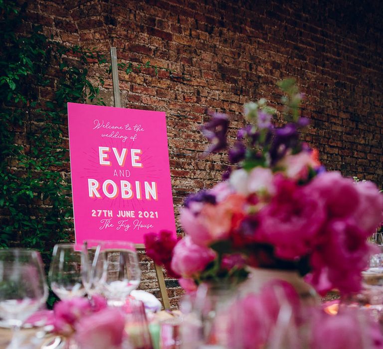 Neutral wedding tablescape with pops of pink, pink floral arrangement with pink peonies, fuchsia roses, lilac sweet peas and green foliage and large pink wedding sign with the couple's names on wooden easel