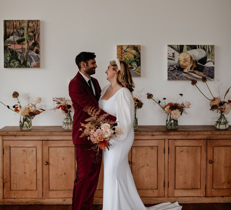 Bride with pretty pearl accessories smiles at the groom in a deep red suit for their autumn wedding at Crumplebury 