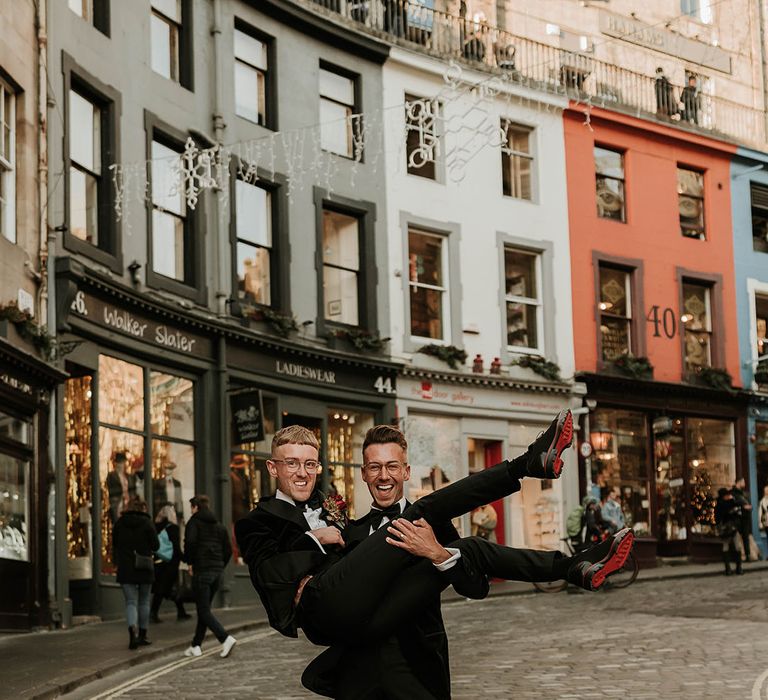 Grooms in black velvet suits, white shirts, black bowties and red rose and foliage boutonnieres with Christian Louboutin groom shoes in the middle of Edinburgh after stylish city wedding