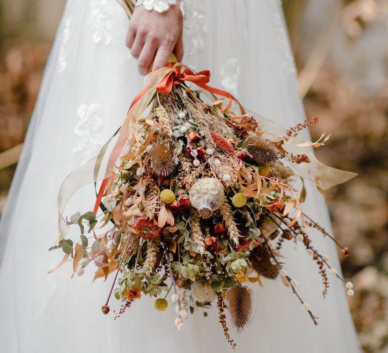 Bride in soft ivory dress with lace floral detailing on the long sheer sleeves holding autumnal floral bouquet with dried flowers, red roses, foliage, pampas grass and orange ribbon