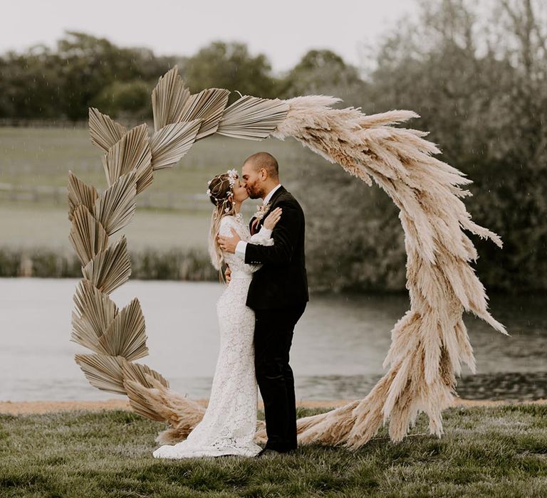 Boho pampas grass moongate and dried palm leaves with the bride and groom sharing a kiss 