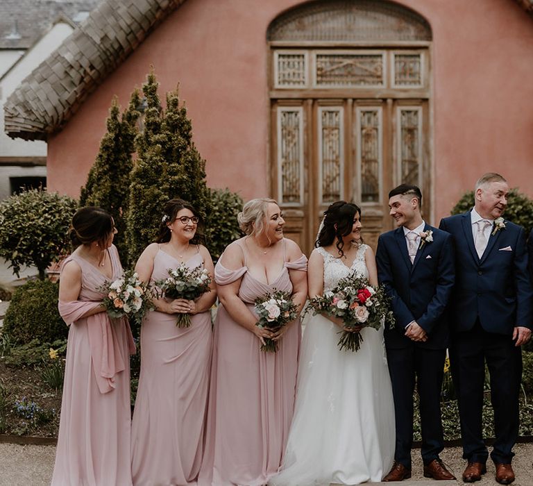 Bride & groom stand beside their wedding party with bridesmaids wearing pale pink bridesmaid dresses in differing styles 