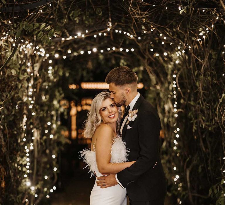 Groom in classic Ralph Lauren black tuxedo with black bowtie and dried flower boutonniere and bride in strapless white satin look feather jumpsuit embracing in tunnel of foliage and fairy lights at The Old Kent Barn wedding venue