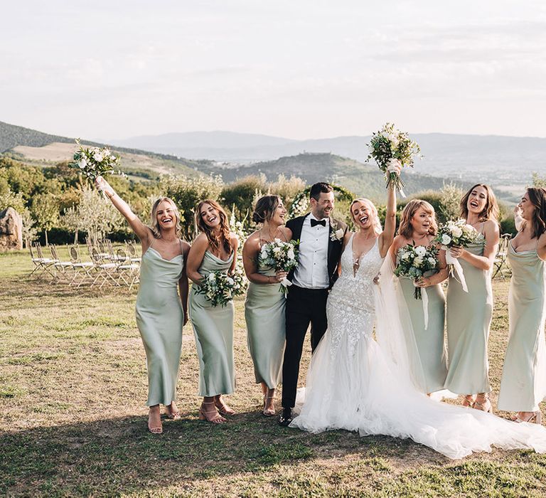 Bride in lace wedding dress beside her groom in black tie and bridesmaids in satin cowl neck bridesmaid dresses