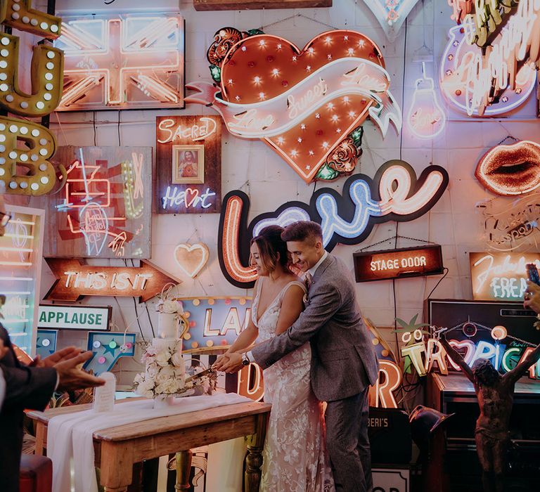 Bride and groom cutting the three tiered buttercream wedding cake amidst a plethora of colourful vibrant neon wedding signs and LED wedding signs 