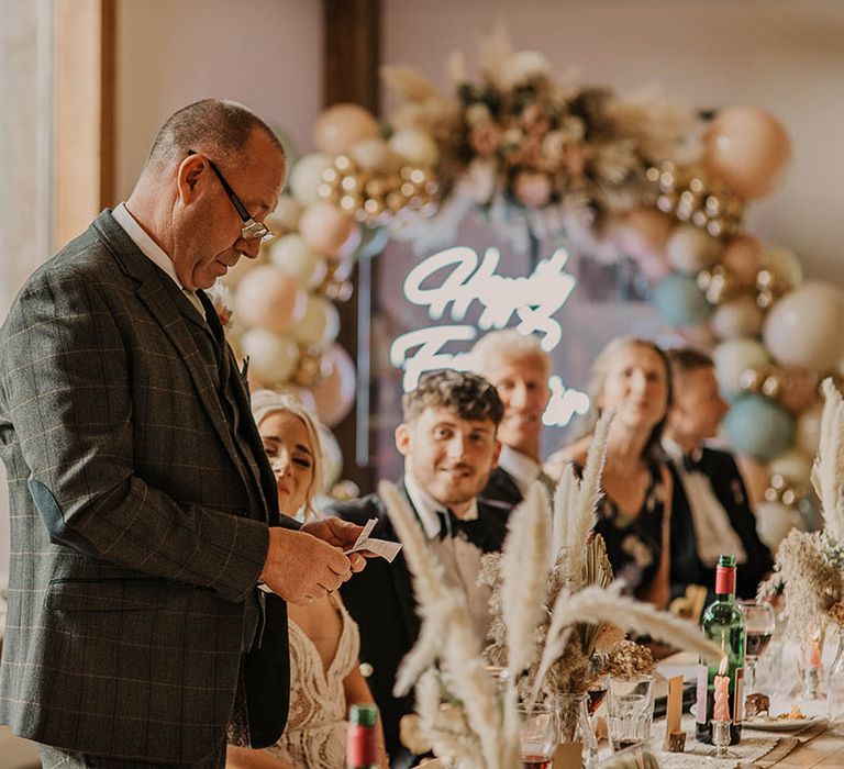 Father of the bride in three piece grey checkered suit wears glasses as he reads his wedding speech at rustic barn wedding with pampas grass, neon sign and balloon arch decorations 