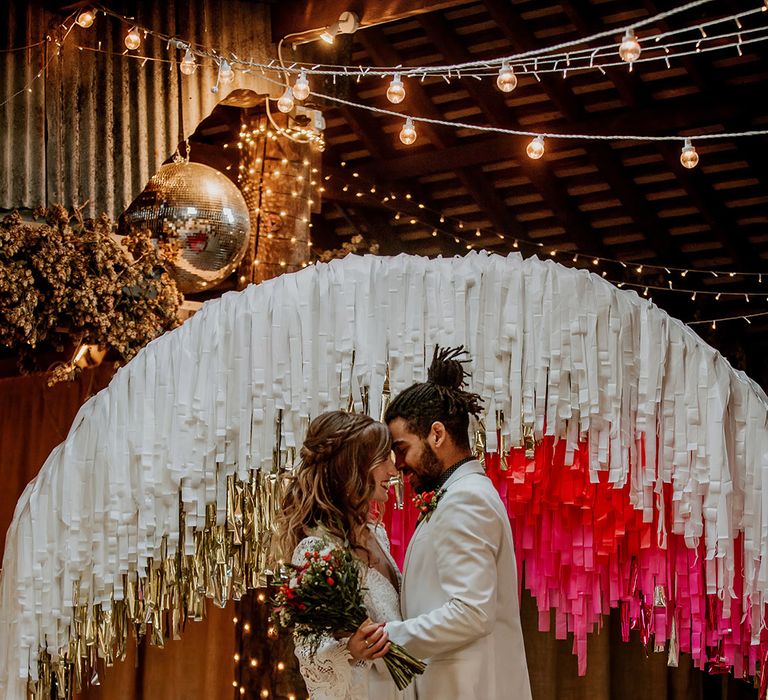 Bride in long sleeve boho wedding dress and groom in white blazer with black suit trousers embracing at the aisle with fairy lights, disco ball decorations, and large semi-circle arch decor with white, gold and hot pink streamers 