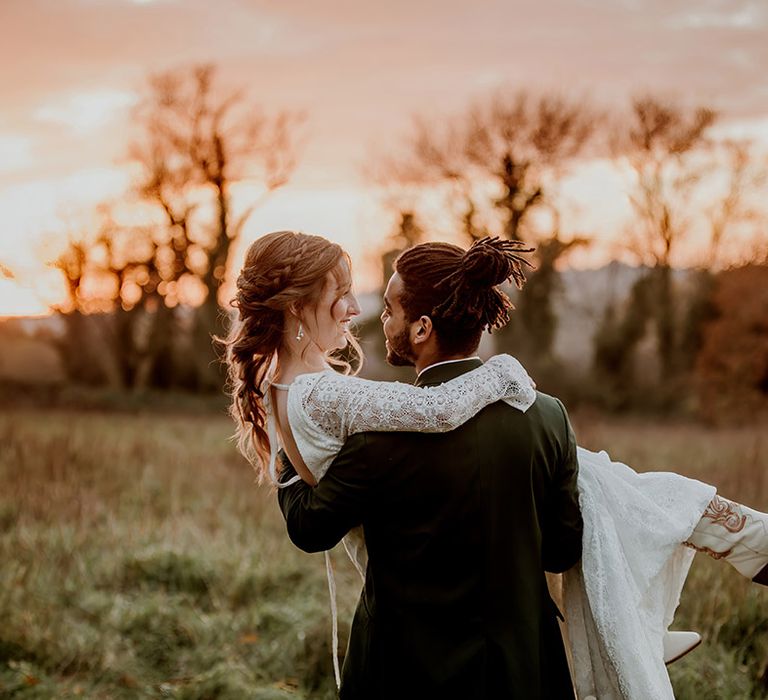 Bride with plait hair styling wearing long sleeve lace wedding dress with open-back detail being carried by groom in black tux at golden hour 