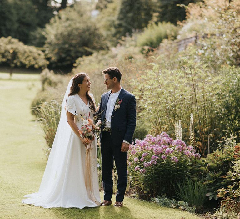 Bride in embroidered handmade wedding dress with groom in navy suit enjoying the summer sun 