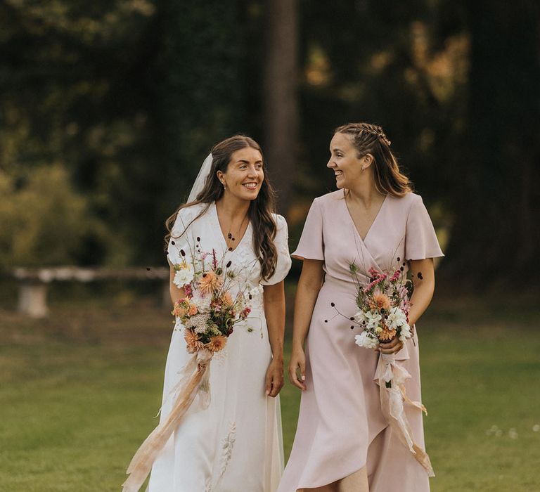 Bridesmaid in pink wrap bridesmaid dress walking along with the bride in embroidered wedding dress with rustic wedding flowers 