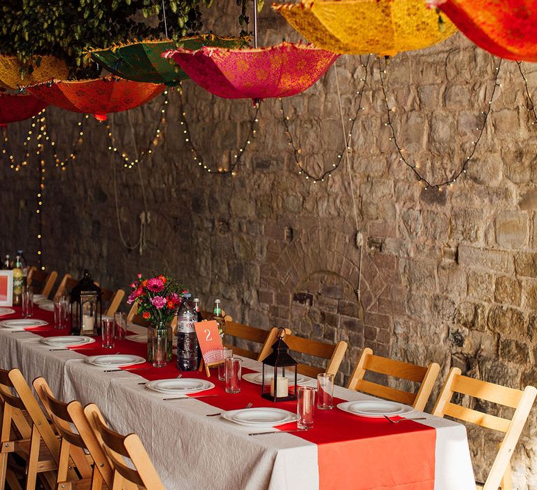 Pink, yellow and orange Indian umbrellas hang above colourful table complete with orange table runner and colourful floral arrangements 