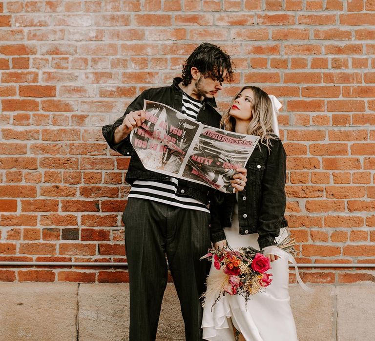 Bride in personalised black denim jacket and satin wedding dress looking up at groom in black and white striped shirt, grey pinstripe trousers, Dr Martens boots and black personalised denim jacket reading tabloid-style wedding stationery 