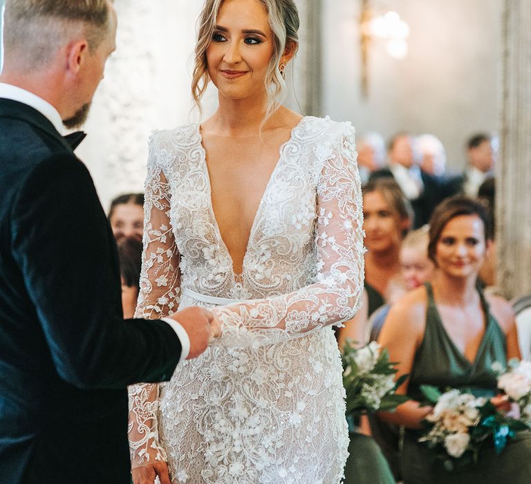 Bride in intricate floral 3D lace wedding dress with plunging neckline and shoulder pads standing at the altar with the groom 