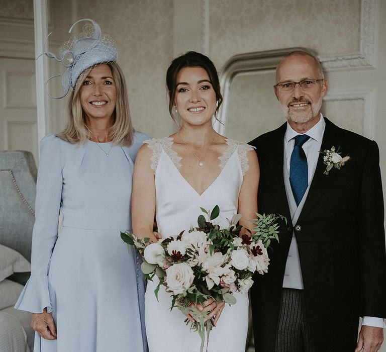 Bride wears Savannah Miller wedding dress and holds classic bridal bouquet whilst stood beside her parents 