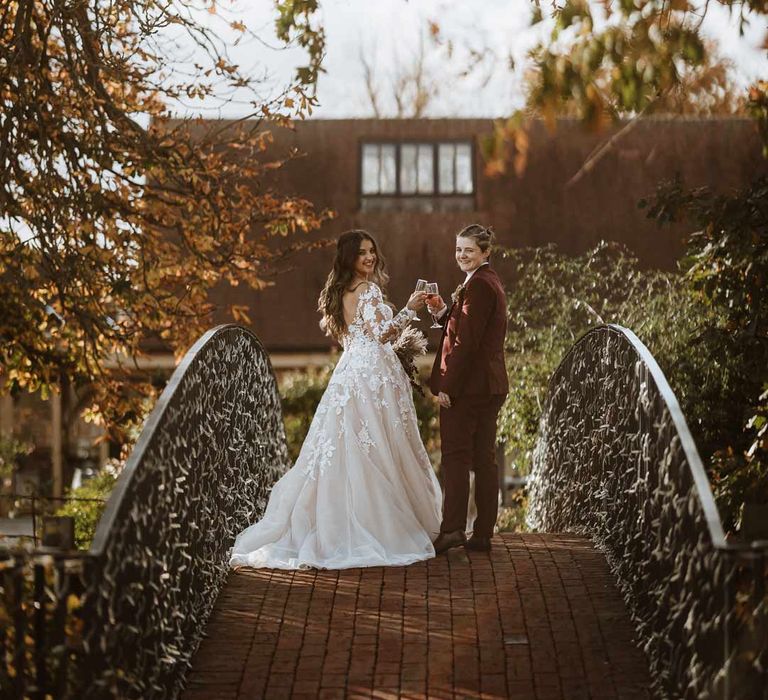 Bride in maroon bridal suit with grey waistcoat on bridge at Bassmead Manor Barns wedding venue with bride in long sleeve lace illusion sleeve wedding dress with overlay 