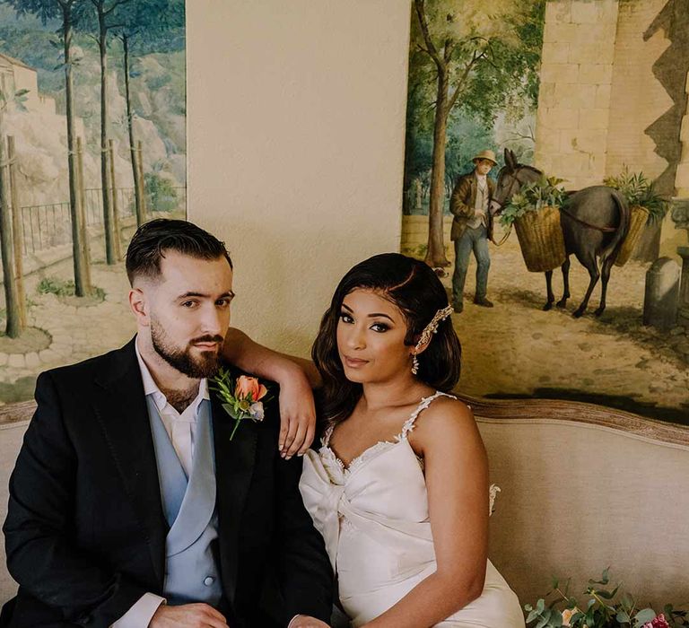 Groom in black blazer with pink garden rose and foliage boutonniere, cornflower blue waistcoat and bride in bow front sleeveless satin wedding dress with large gold bridal hair clip 