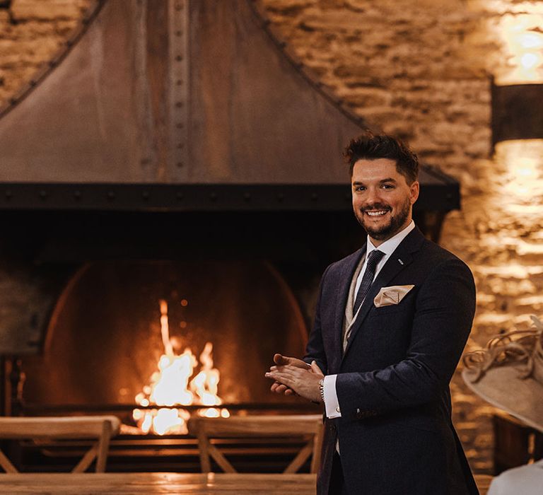 Groom smiles brightly wearing a navy suit and spotted tie with a cream waist coat on waiting for the bride at the end of the aisle 