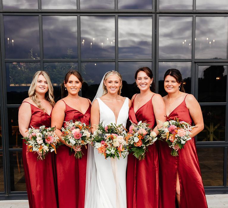 Bridesmaids in matching red satin bridesmaid dresses with leg slit posing with the bride with her curled blonde hair in sleek hairstyle with fitted wedding dress 