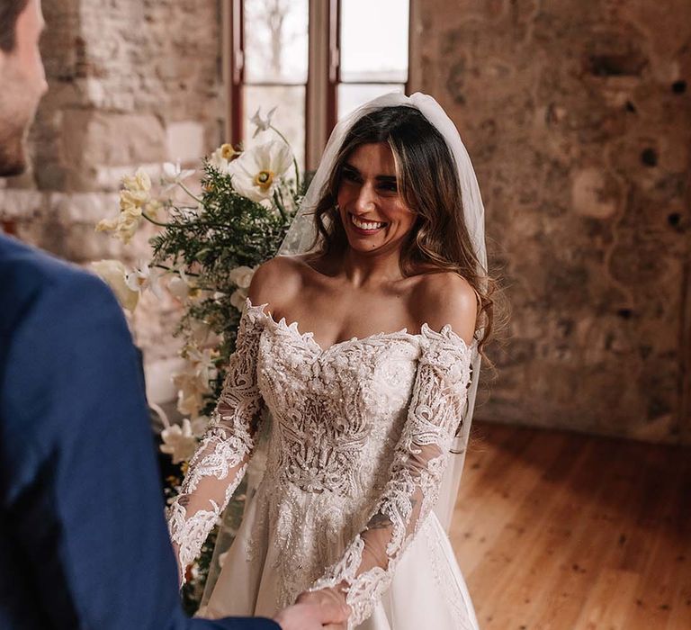 Bride in sequinned off the shoulder wedding dress smiling at the altar holding hands with her groom 