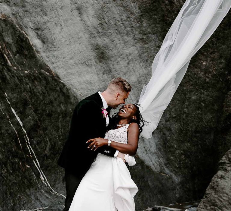 Groom in bottle green velvet grooms blazer holding bride in v neck sleeveless lace Pronovias wedding dress, cathedral length veil and silver sparkly bridal jewellery on Tunnels Beaches 