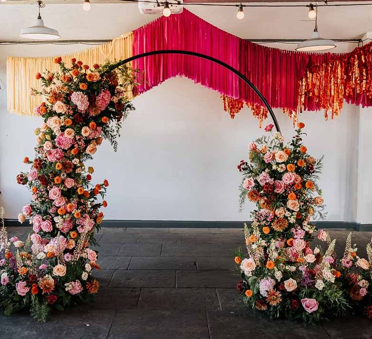 Large circular floral arch with vibrant flowers and foliage attached and pink, red, yellow and metallic wedding streamers behind at reception room of Northern Monk Refectory Leeds 