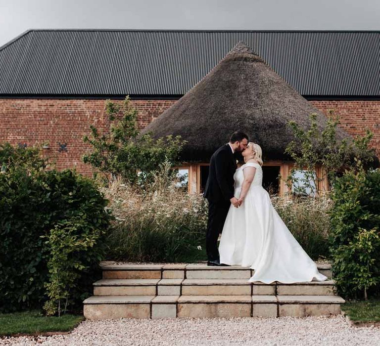 Bride in satin off the shoulder wedding dress kissing groom in classic black grooms three piece tuxedo with white rose boutonniere and black bowtie outside of Brickhouse Vineyard Devon wedding venue