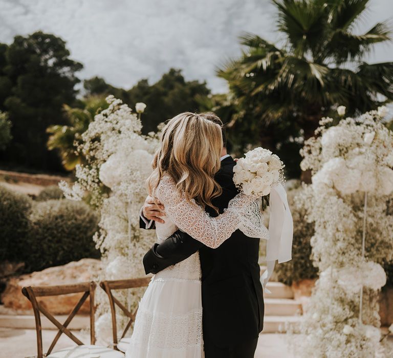 destination wedding with bride in a lace wedding dress embracing her groom in a tuxedo in front of all white column wedding flowers