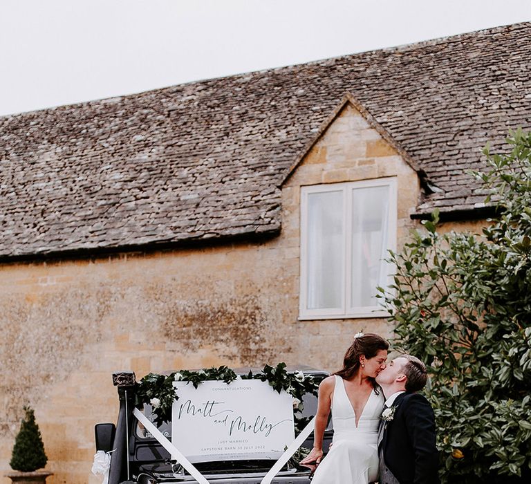 Black Landrover wedding car with white ribbon, flower bumper decorations and white welcome wedding sign with the bride and groom sharing a kiss