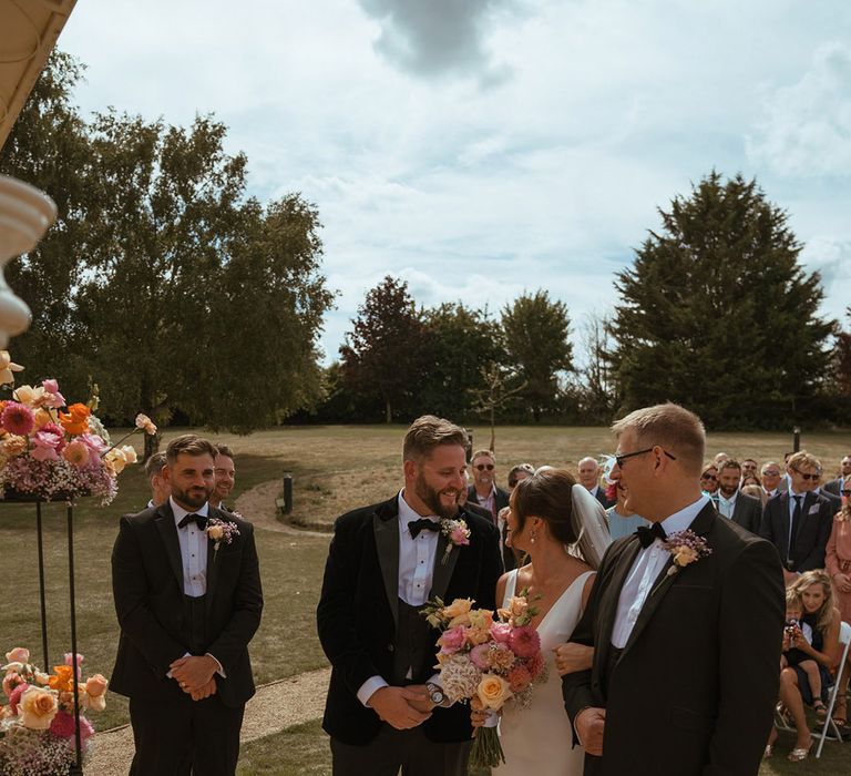 Father of the bride walks the bride to the end of the aisle with the groom and best man standing in black tuxedo with pink and orange rose floral decorations 