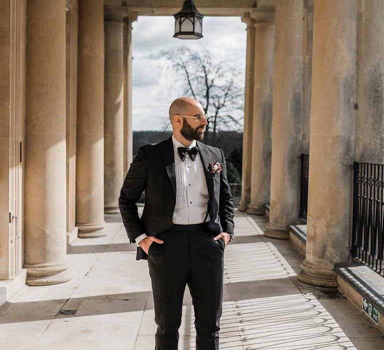 Groom in classic black tuxedo with bowtie and wildflower boutonniere standing on grounds of Buxted Park wedding venue