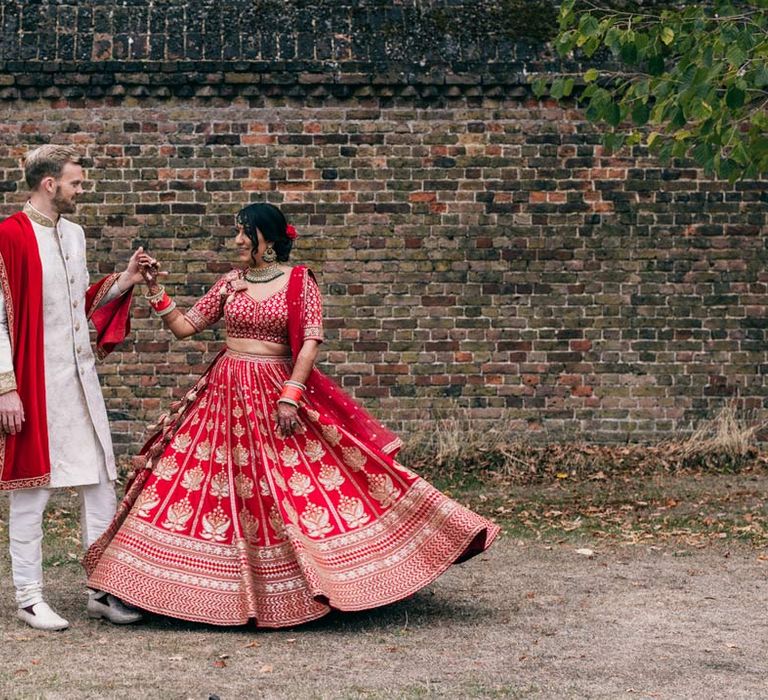 Groom in traditional white and gold sherwani dancing with bride in red and gold lehenga and gold and forest green bridal jewellery on the grounds of Templars Barn