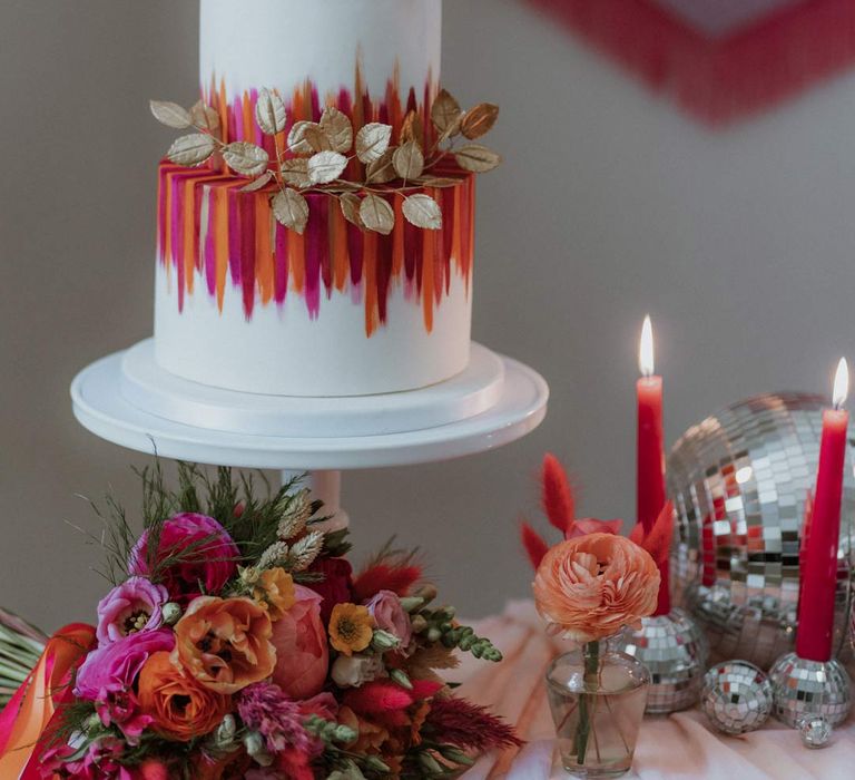 Three tiered iced wedding cake with pink and orange icing stripes and gold decorative leaves as cake toppers on table with dusky pink tablecloth, pink and orange streamers and disco ball wedding decor