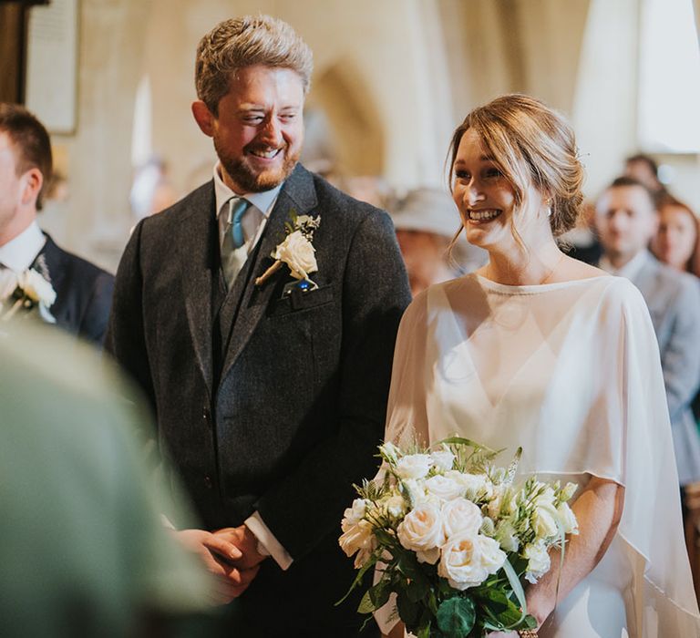 Bride wearing simple white wedding dress with sheer overlay standing with the groom at the aisle 