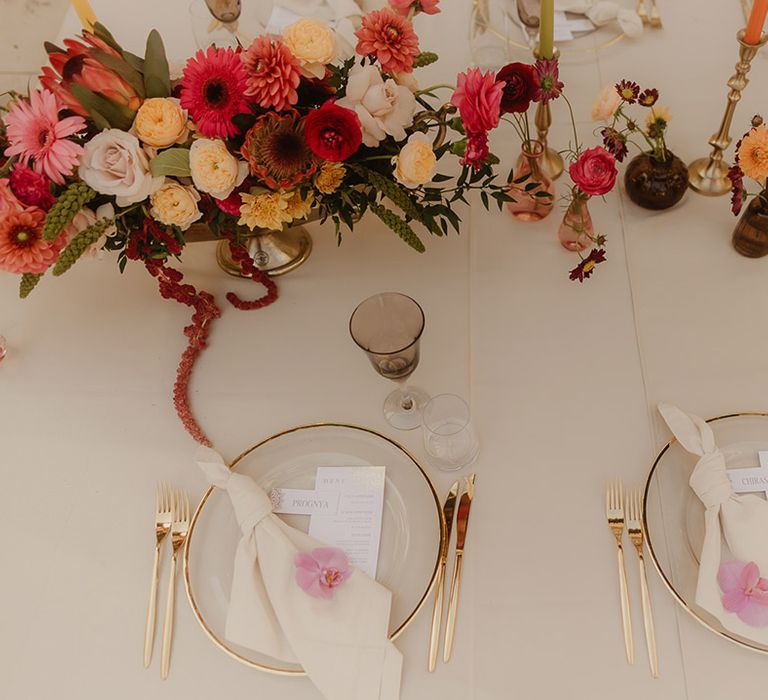 place settings with gold rim charger plate tableware and cutlery, and brightly coloured flowers and candle centrepiece decor 