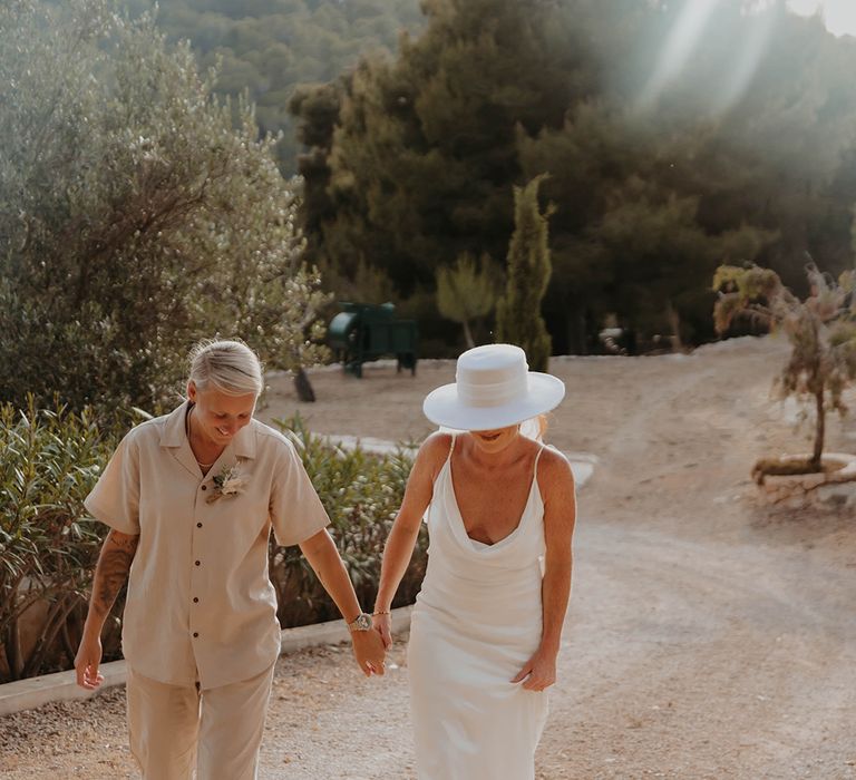 Bride in beige short and trousers coordinates walking with the bride in a white satin dress and bridal hat 