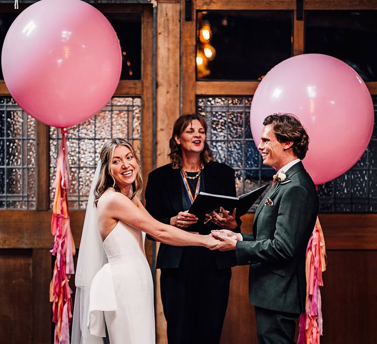 The bride and groom hold hands for their wedding ceremony with large pink wedding balloons and streamers decorating the altar space 