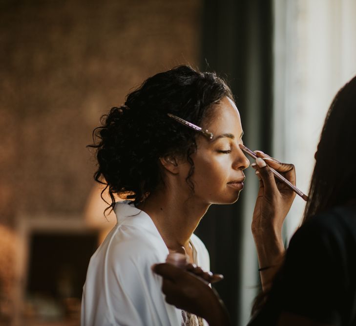 A Black bride with curly low bun is having her makeup applied.