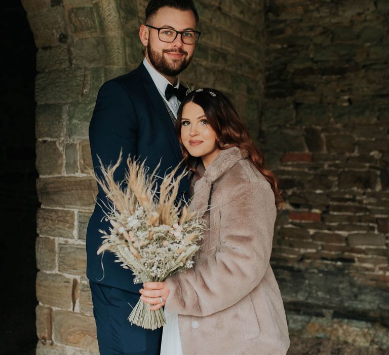 Bride in fur coat holding dried flower bouquet hugs groom in stone building