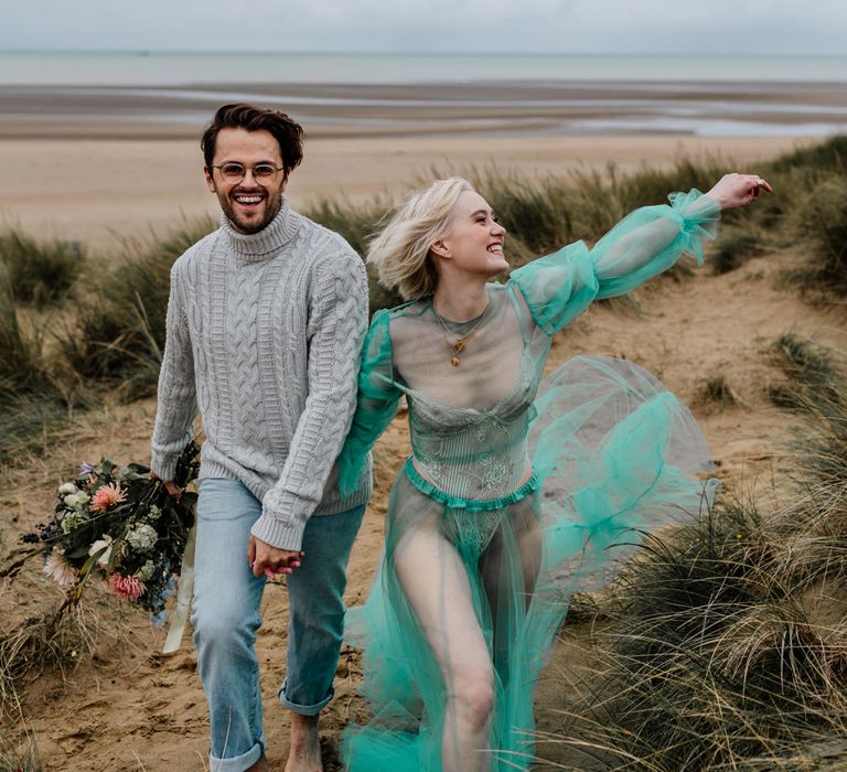 White brunette bearded groom in glasses, grey jumper and faded jeans walking through the sand dunes with his blonde bride in a sheer green pastel wedding dress and body suit