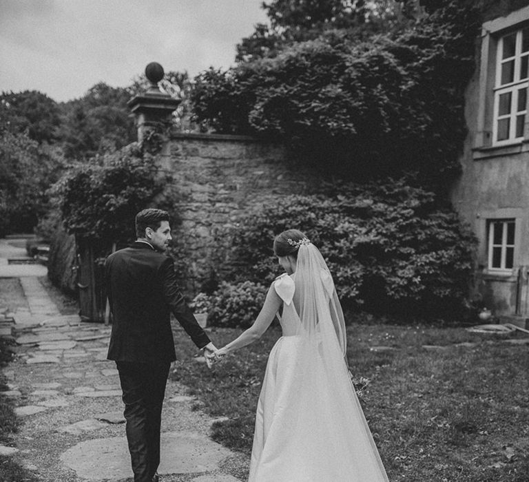 Bride in white Jesus Piero gown and cathedral length veil holds hands with groom in black Tiger of Sweden tux in gardens at German wedding