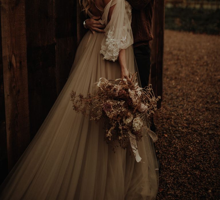 Groom in a wooly jumper kissing his bride in a floaty wedding dress as she holds a dried flower wedding bouquet 