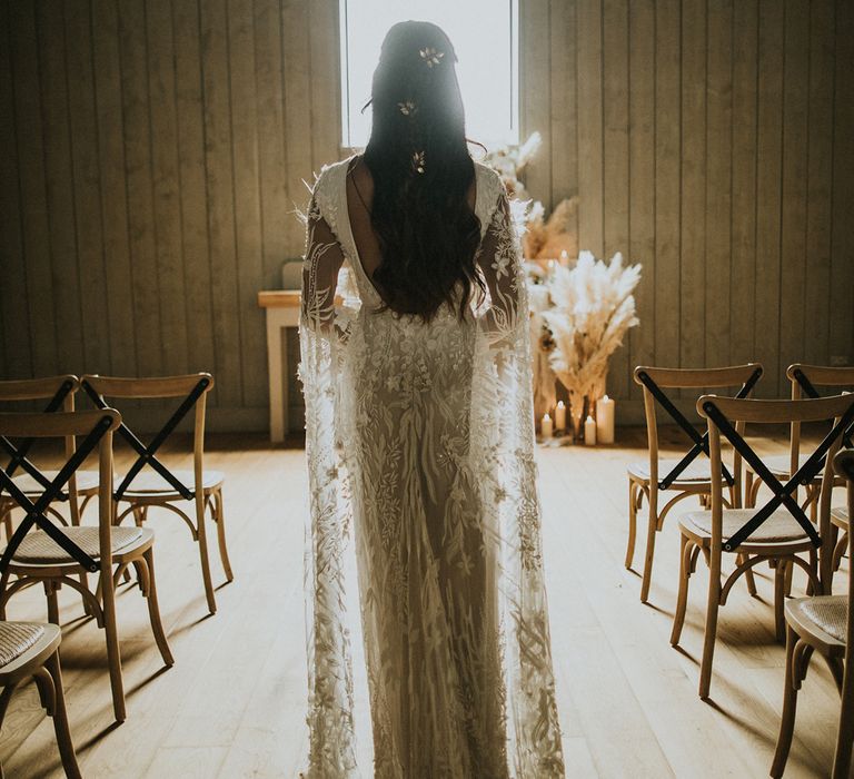 Boho bride in a lace wedding dress standing in the ceremony room with light flooding in the window 